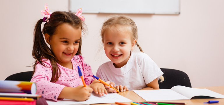Two young children sitting at a table, drawing in notebooks with coloured pens, with art supplies spread out in front of them.