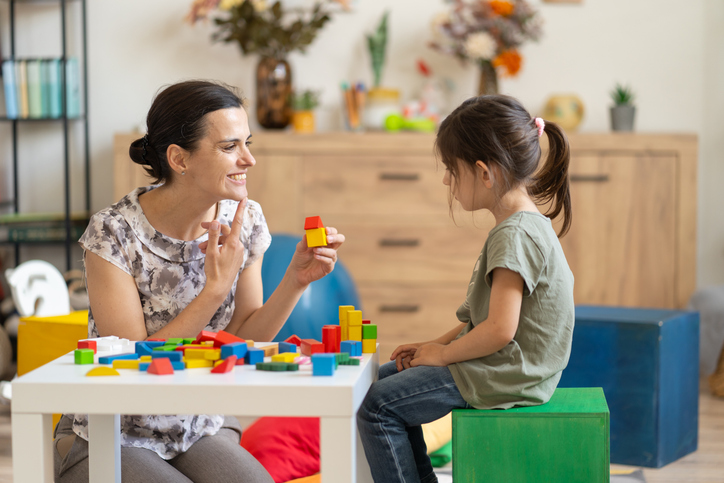 Two people, one adult and one child, are sitting at a table playing with colorful building blocks. They appear engaged and happy as they interact with each other. The background features a tidy room with a wooden shelf.