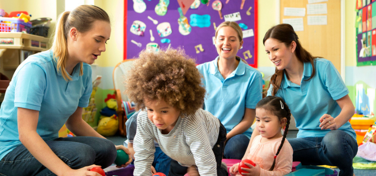 Three childcare workers wearing blue shirts are interacting with young children in a colorful classroom decorated with children's artwork.